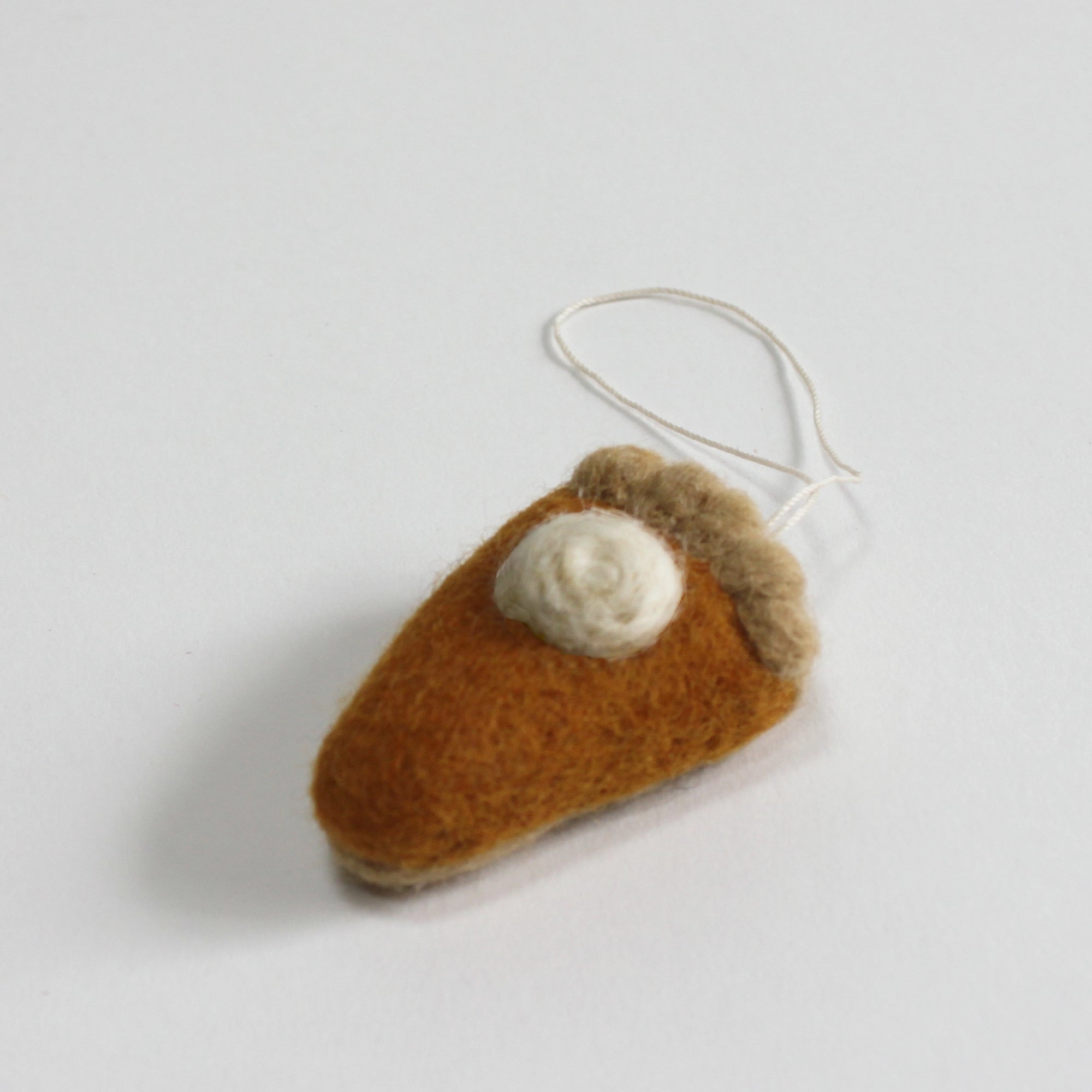 The Faire Felted Ornament is a handmade pumpkin pie slice crafted from wool, featuring a dollop of white cream and an attached string loop, displayed on a white surface.