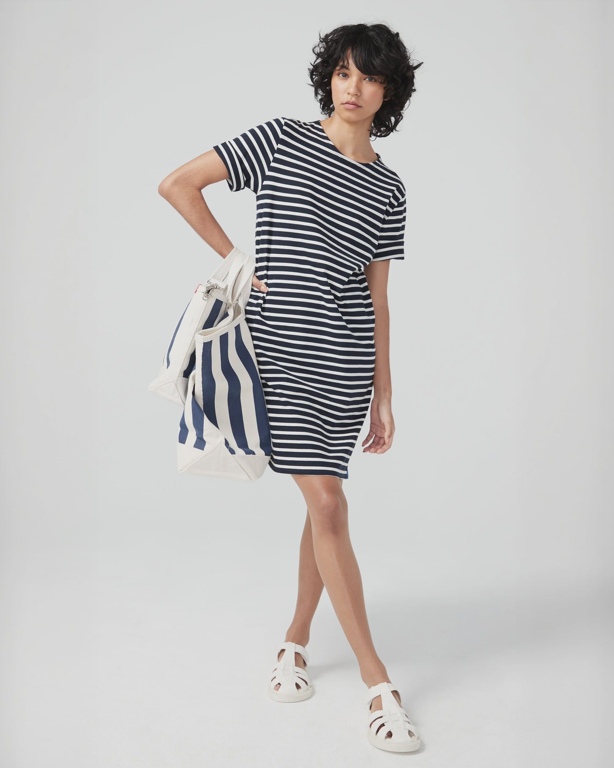 A person with short curly hair stands against a plain light background, wearing a black and white striped dress and white sandals, holding Kule’s The All Over Striped Tote.