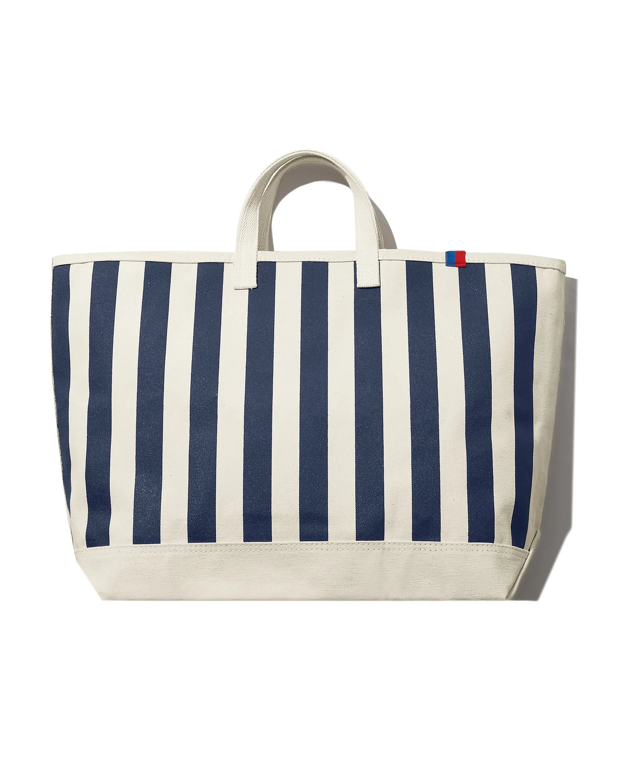 The All Over Striped Tote by Kule is a large cotton canvas bag with vertical navy blue and white stripes, two short handles, and a small red and blue tag at the top right corner. Photographed against a white background.