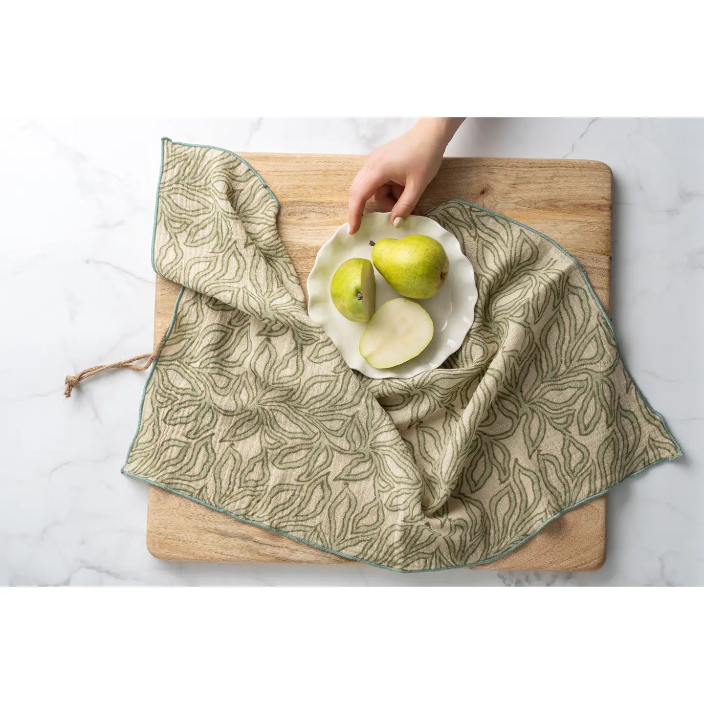A hand places a white plate with two whole green pears and two pear slices on Indaba’s Nori Block Print Napkins atop a wooden cutting board, all set on a white marble surface.