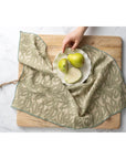 A hand places a white plate with two whole green pears and two pear slices on Indaba’s Nori Block Print Napkins atop a wooden cutting board, all set on a white marble surface.