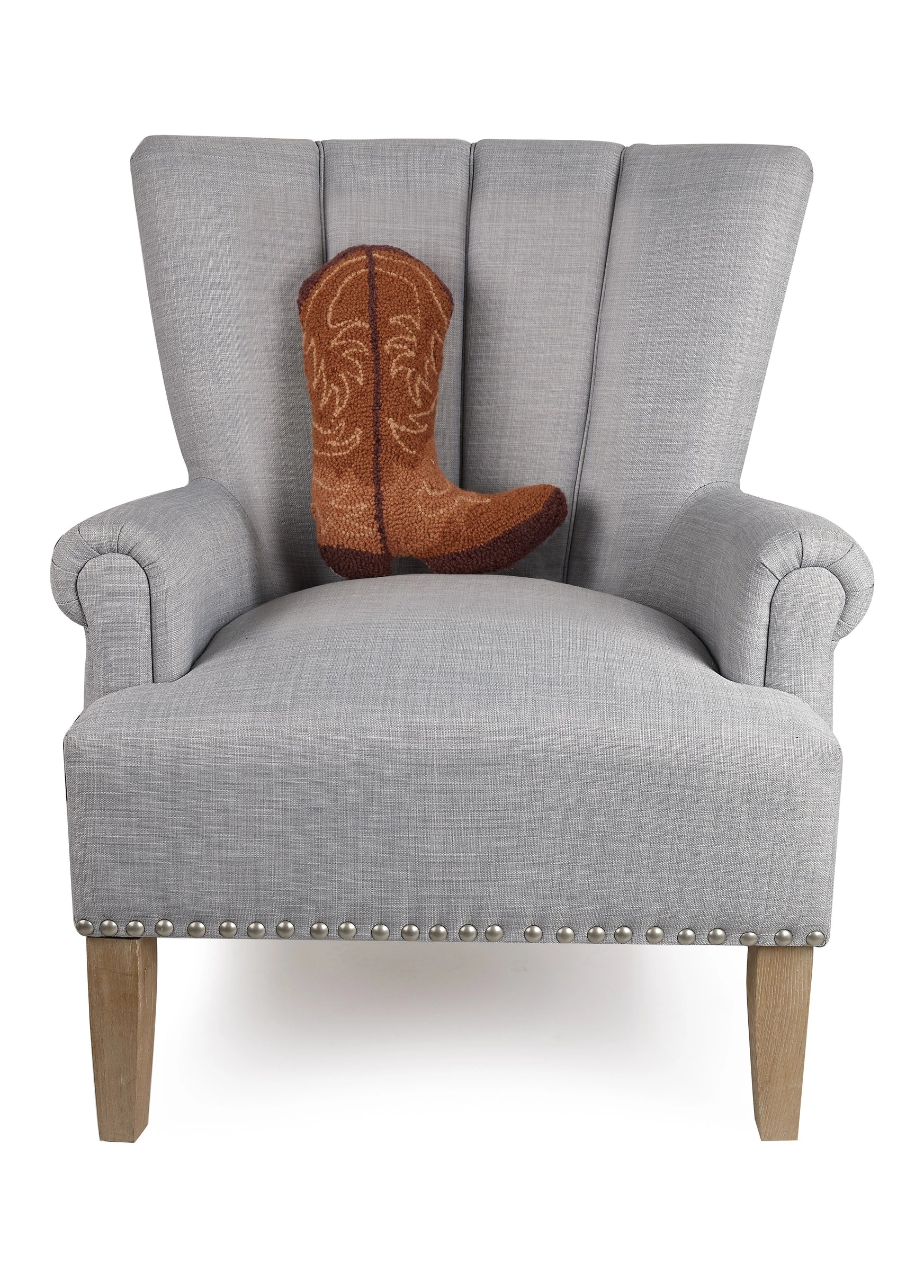 A light gray upholstered armchair with wooden legs and silver studs features the Faire Brown Cowboy Boot Pillow, a poly velvet throw pillow shaped like a brown cowboy boot, placed on the seat.