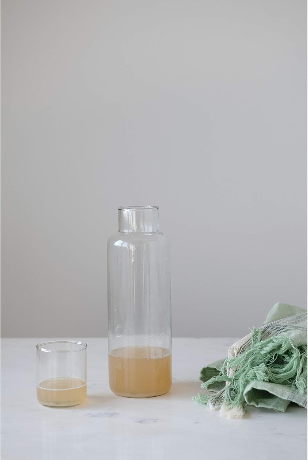 The Creative Co-op Decanter With Glass, partially filled with a light brown liquid, rests on a white surface beside a folded green fringed cloth—elegant tableware for any setting against a plain light gray background.