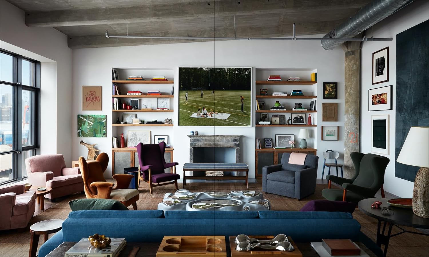 A stylish living room showcases eclectic furniture, a blue sofa, and art, as seen in "Robert Stilin New Work" by Hachette Book Group. Expansive windows fill the space with light and bookshelves display striking artwork above the fireplace.