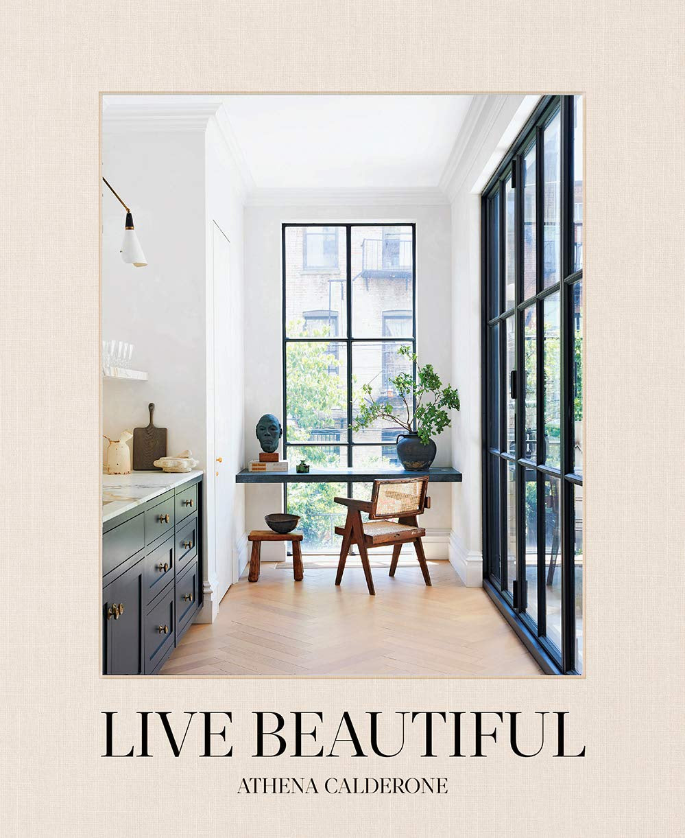 The cover of "Live Beautiful" by Athena Calderone, published by Random House (ISBN: 978141742804), features a serene interior with black-framed windows, a small wooden table, lush plants, glimpse of dark kitchen cabinets against a beige textured backdrop.