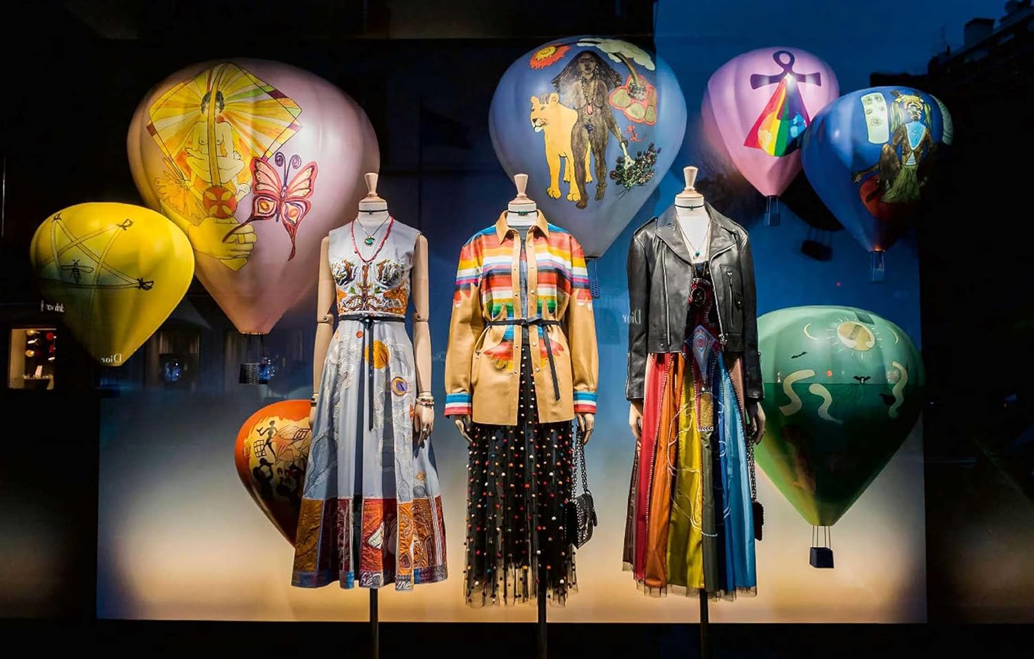 Three mannequins display “Paris In Fashion” by Simon & Schuster Inc., wearing vibrant, artistic outfits inspired by Paris, in front of painted hot air balloons and a dark backdrop in a stylish window display.