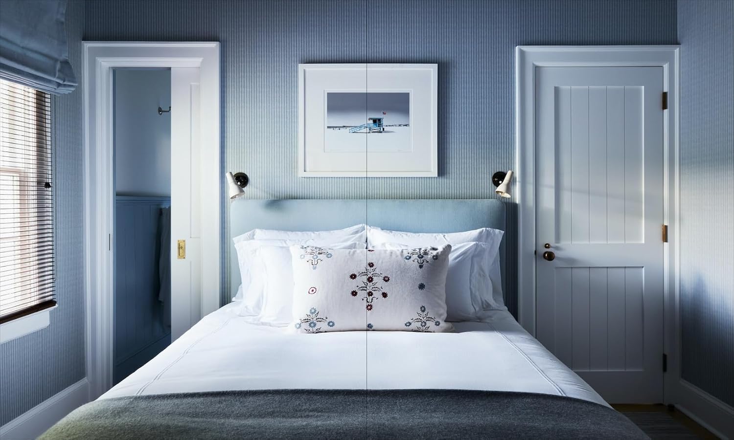 A bed with white linens and embroidered pillows evokes the style of Robert Stilin New Work by Hachette Book Group, centered in a blue-walled room between two doors, sunlight streaming through blinds onto reading lights and art above the bed.