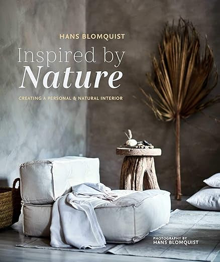 A cozy interior setting with soft white cushions, a natural wooden stool, and a large dried leaf decoration against a textured gray wall. The cover text reads "Inspired By Nature" by Random House—a perfect guide for cultivating emotional well-being.