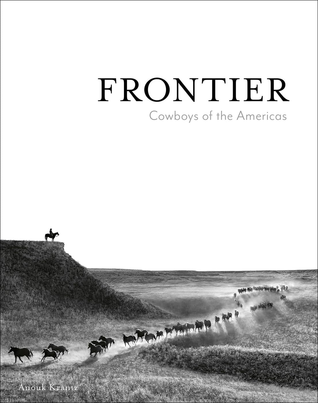 Cover of "Frontier Cowboys of the Americas" by National Book Network features a striking B&W photo of a cowboy on horseback on a hill, capturing American ranch culture as he oversees horses moving through the grassy landscape below.