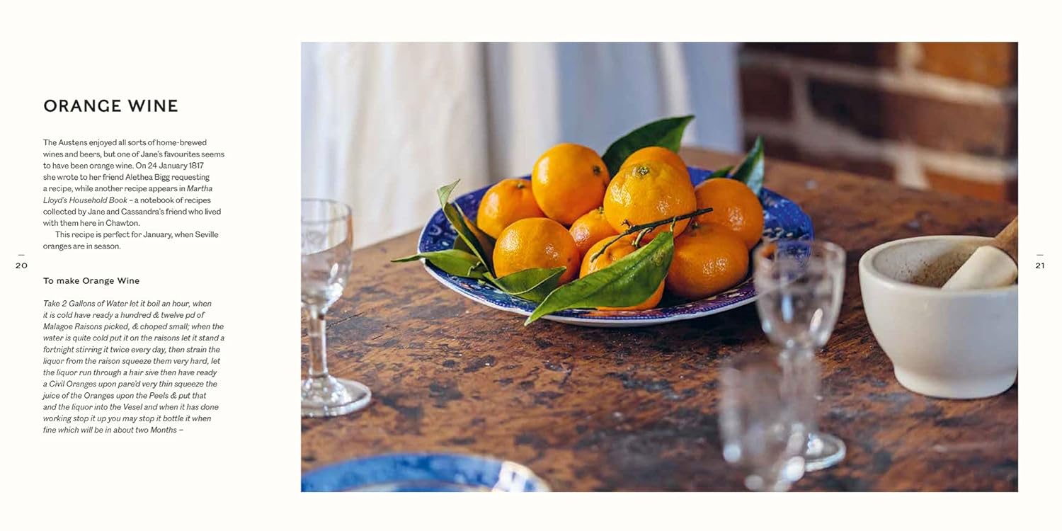 A blue plate with whole oranges and green leaves sits on a rustic wooden table amid clear glasses and a brick wall, evoking an atmosphere reminiscent of "A Jane Austen Year" from Random House.