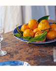 A blue plate with whole oranges and green leaves sits on a rustic wooden table amid clear glasses and a brick wall, evoking an atmosphere reminiscent of "A Jane Austen Year" from Random House.