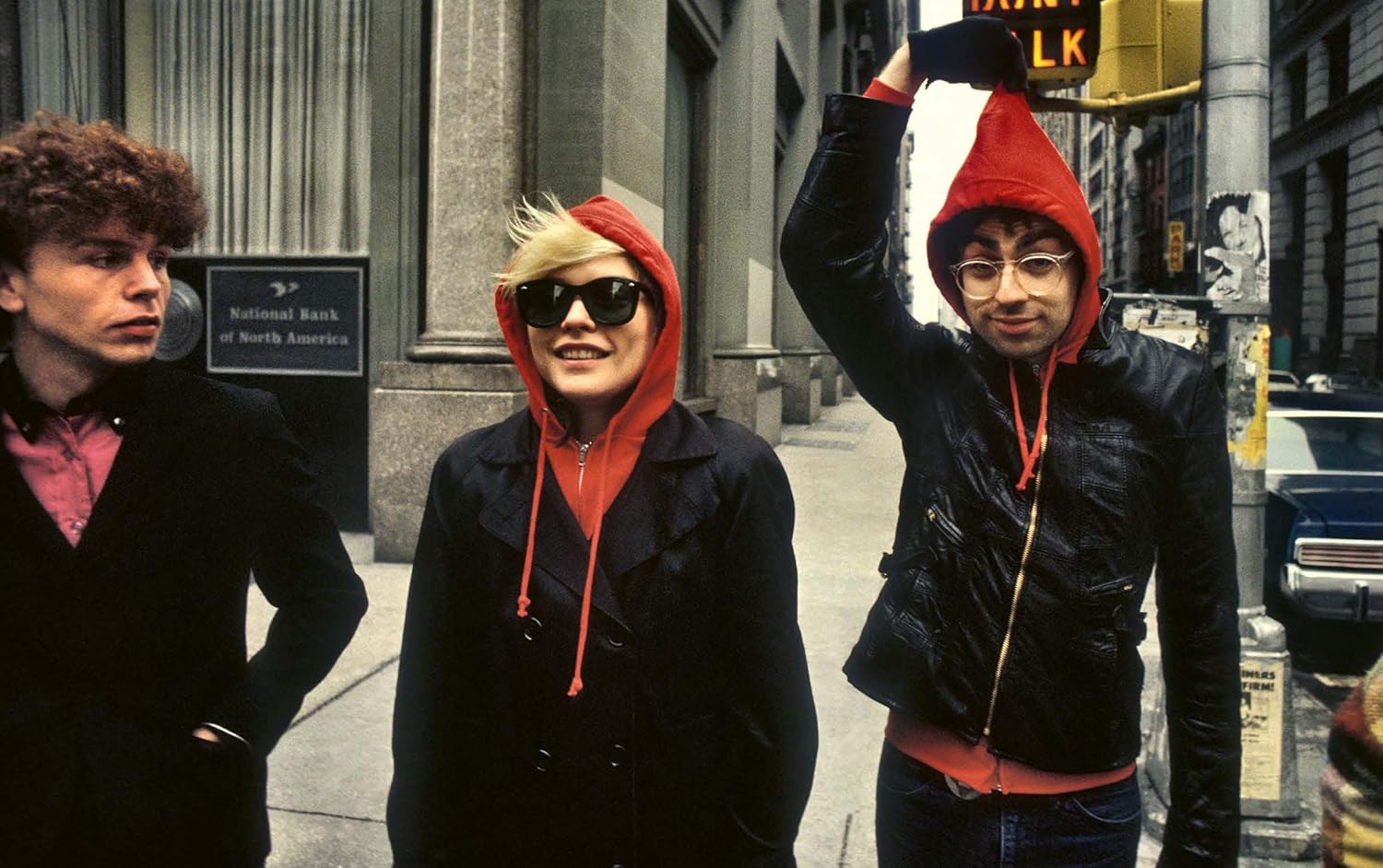 Three people walk on a city sidewalk. One, in sunglasses and a red hoodie under a black jacket, channels Blondie’s style as seen in Martyn Goddard's photos from "Blondie In Camera 1978" by Simon & Schuster Inc., with urban buildings behind them.
