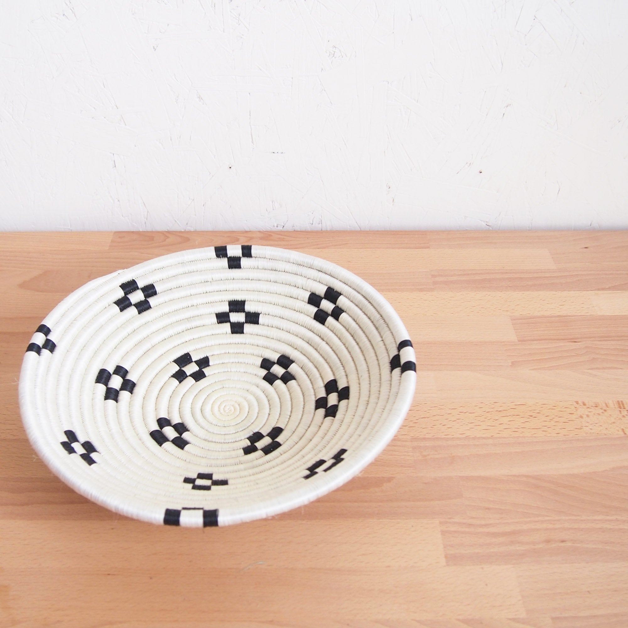 The Maraba Small Bowl by Amsha - Faire, a round sisal basket with a white base and black geometric patterns, rests on a light wooden surface against a textured white wall.