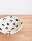 The Maraba Small Bowl by Amsha - Faire, a round sisal basket with a white base and black geometric patterns, rests on a light wooden surface against a textured white wall.