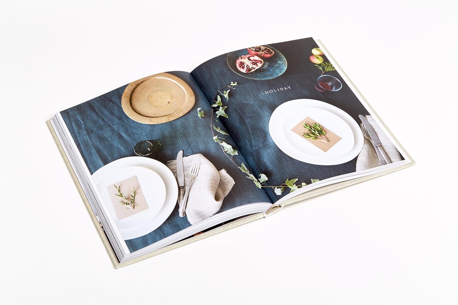 The book "Sunday Suppers" by Random House is open to a scene of two white plates, beige napkins, greenery on a dark blue cloth, and a wooden bowl with fruit—an inviting table set for communal cooking and gathering friends.