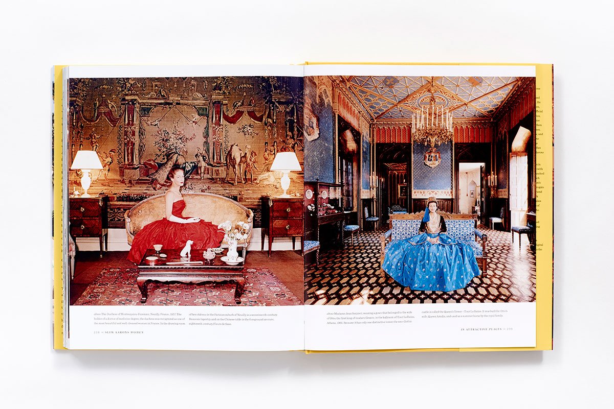 An open copy of "Slim Aarons: Women" by Hachette Book Group displays photos of extraordinary women in glamorous ball gowns—one in red on the left, one in blue on the right—channeling iconic high-society style.