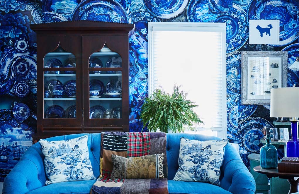 A living room inspired by Sean Scherer's Kabinett from Hachette Book Group features blue patterned wallpaper, a blue sofa with patterned pillows, vintage blue dishes in a wooden cabinet, a potted fern, and framed art by a bright window.