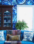 A living room inspired by Sean Scherer's Kabinett from Hachette Book Group features blue patterned wallpaper, a blue sofa with patterned pillows, vintage blue dishes in a wooden cabinet, a potted fern, and framed art by a bright window.