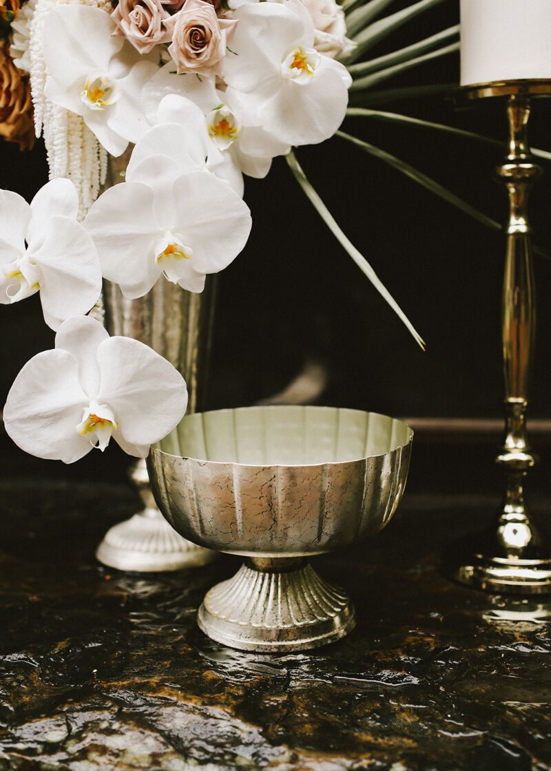 The Accent Decor Lita Compote L in antiqued silver rests on a dark marble surface beside a tall vase of white orchids and roses, with a gold candlestick positioned in the background.