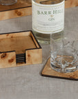 A bottle of Barr Hill gin, an antler-design glass, and a Zodax Valberg Burl Coaster rest on beige fabric. A stack of matching coasters is neatly organized in the Valberg Burl coaster holder nearby.