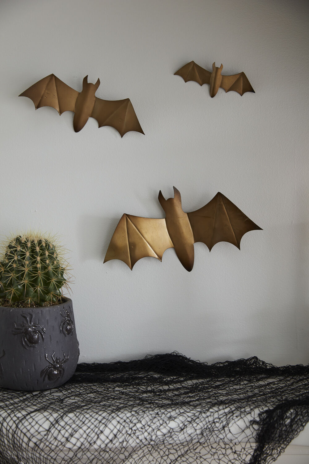 Three Bat Wall Art accents by Accent Decor are mounted on a light wall above a table with a dark cloth and a potted cactus in a gray planter, adding the perfect spooky touch to your home decor.
