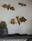 Three Bat Wall Art accents by Accent Decor are mounted on a light wall above a table with a dark cloth and a potted cactus in a gray planter, adding the perfect spooky touch to your home decor.