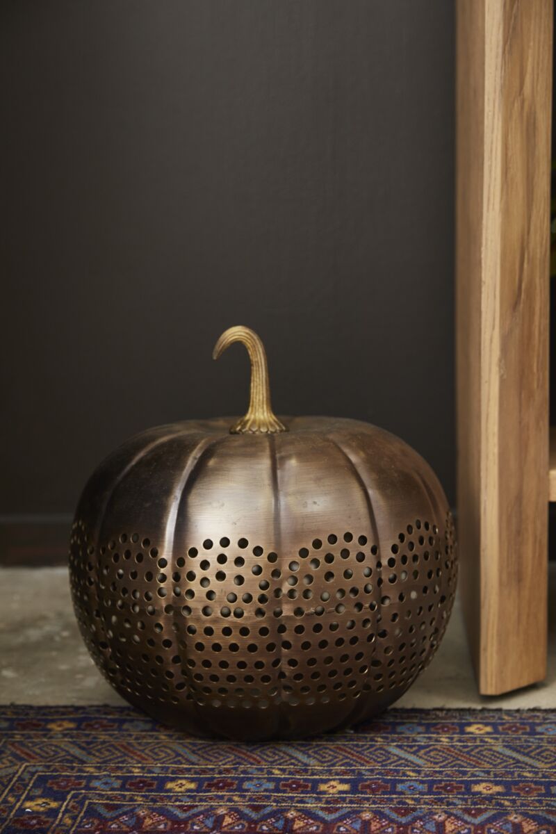 The Accent Decor Harvest Pumpkin Lantern 14x14, featuring a perforated sheet metal design, rests on a patterned rug beside a wooden structure against a dark background.