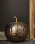 The Accent Decor Harvest Pumpkin Lantern 14x14, featuring a perforated sheet metal design, rests on a patterned rug beside a wooden structure against a dark background.