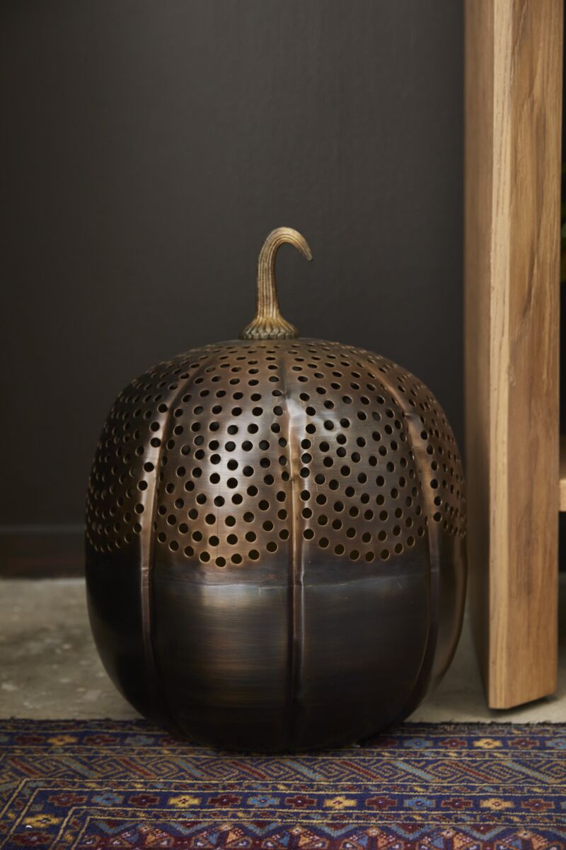 The Accent Decor Harvest Pumpkin Lantern 13x18, with its perforated metal pumpkin design, sits on a patterned rug near wooden furniture against a dark wall, bringing charming fall decor to your porch or living space.