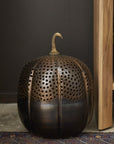 The Accent Decor Harvest Pumpkin Lantern 13x18, with its perforated metal pumpkin design, sits on a patterned rug near wooden furniture against a dark wall, bringing charming fall decor to your porch or living space.