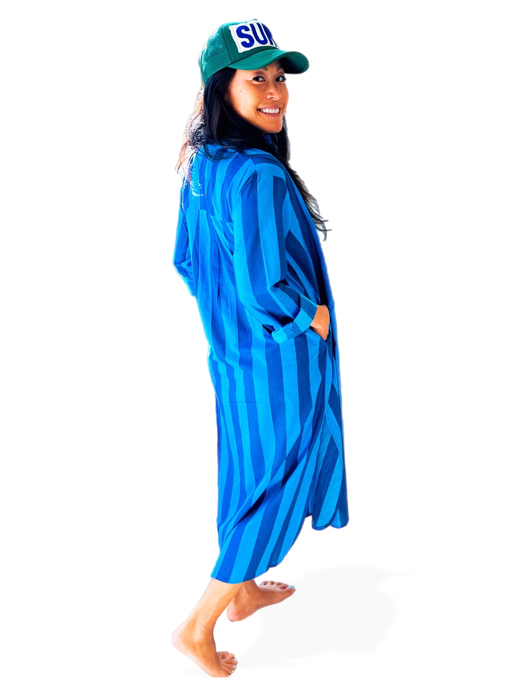A person stands barefoot, smiling over their shoulder, wearing a green "SUN" cap and the Faire Surf Stripe Shirt Dress.