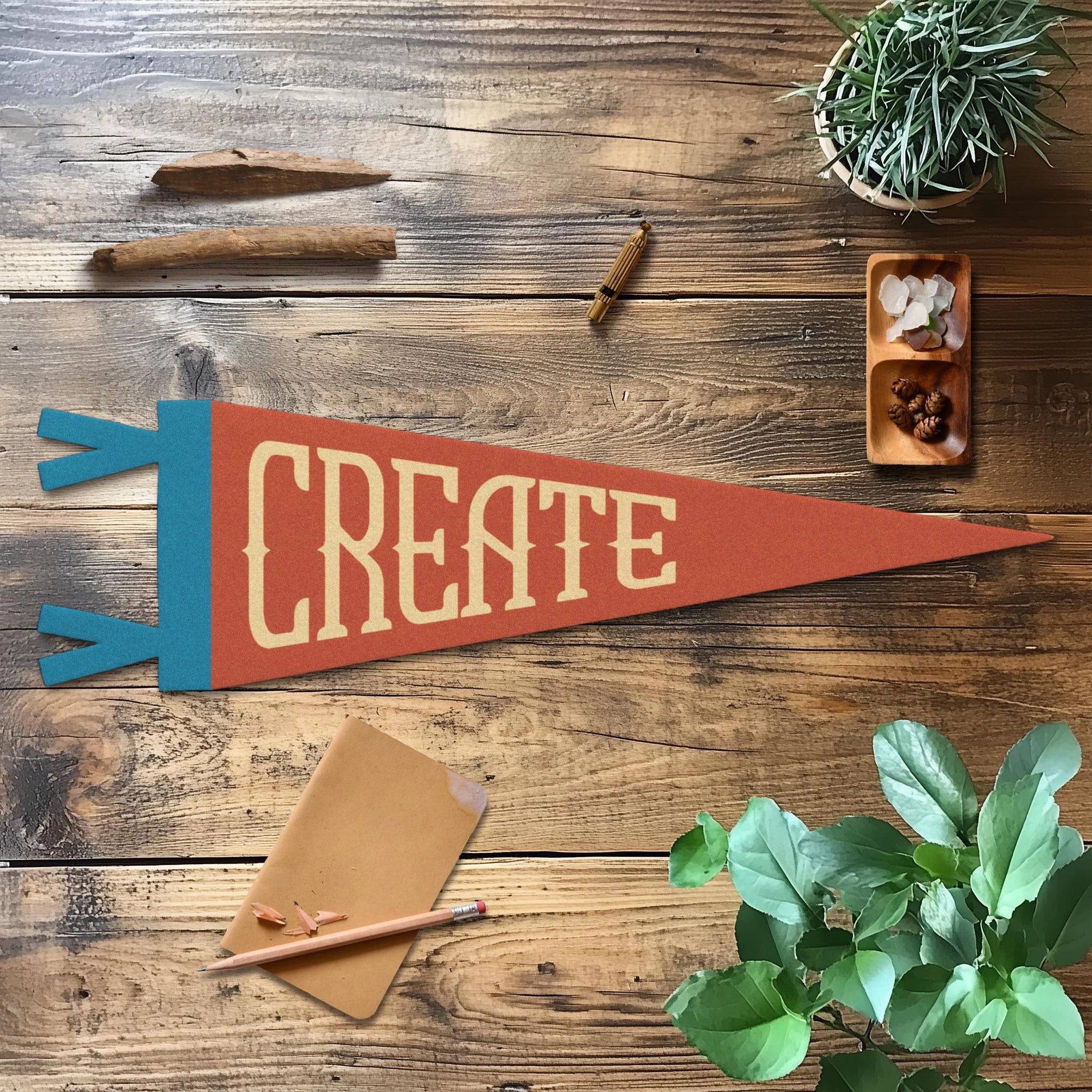 The Faire "Create" Felt Pennant rests on a wooden table with a pencil, brown envelope, cinnamon stick, crystals, and plants—ideal as creative room decor or a thoughtful gift to inspire creativity.