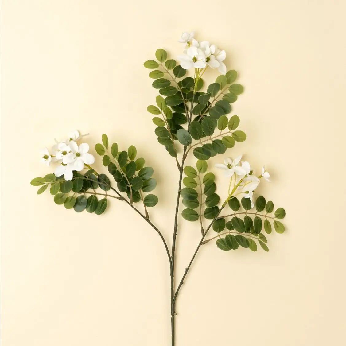 The Indaba Locust Flower Branch features green oval leaves and small white faux flowers, arranged as a decorative accent against a plain light beige background.