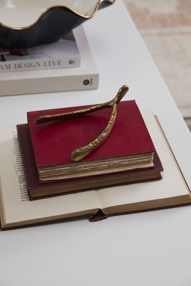 The Wishbone Sculpture by Accent Decor sits atop two stacked red hardcover books on an open book, all placed on a white surface. A white book and a decorative bowl are partially visible nearby.