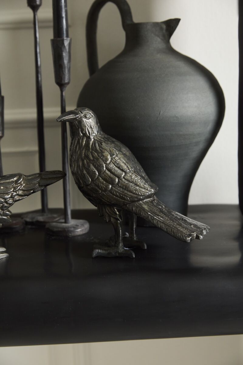 The Nevermore Figurine by Accent Decor stands on a dark shelf before a matte black ceramic pitcher, surrounded by other dark-toned decor for a subtle haunted vibe.