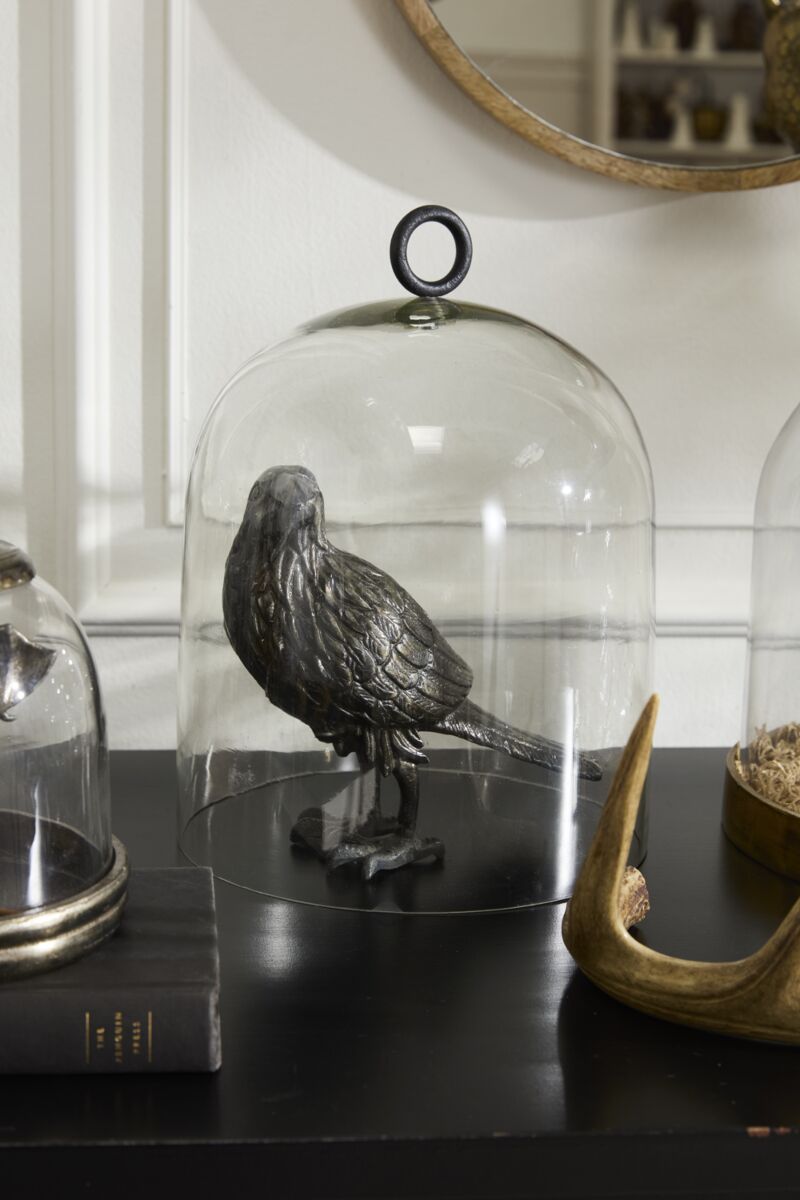The Accent Decor Nevermore Figurine, a metallic raven under glass, sits beside a book and antler on a black surface for an eerie Halloween vibe. A smaller dome with moss adds to the haunting display.