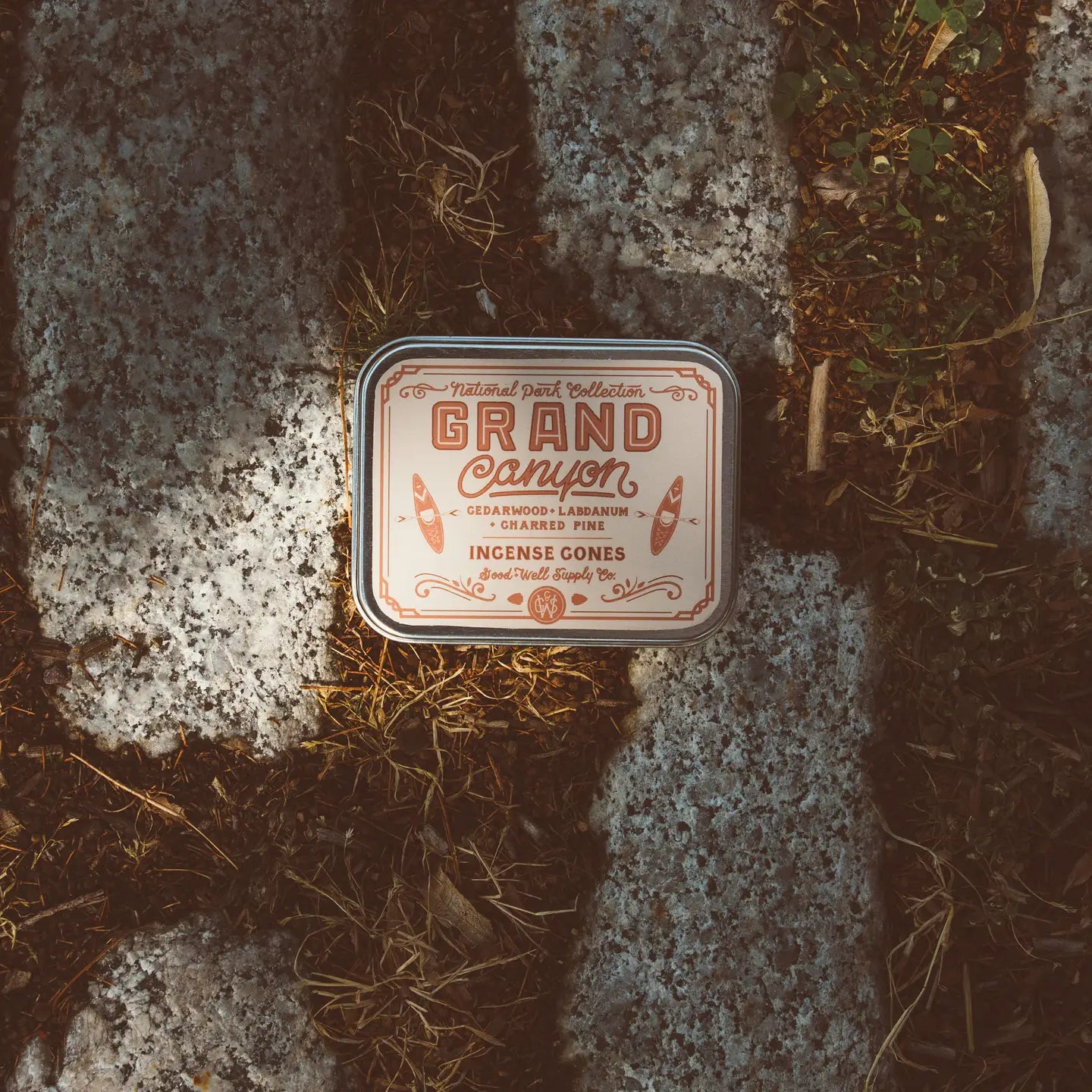A tin labeled "G&W Incense" by Faire sits among brown grass and gray stones, with sunlight and shadows highlighting the scene, evoking the earthy scent of cedarwood.