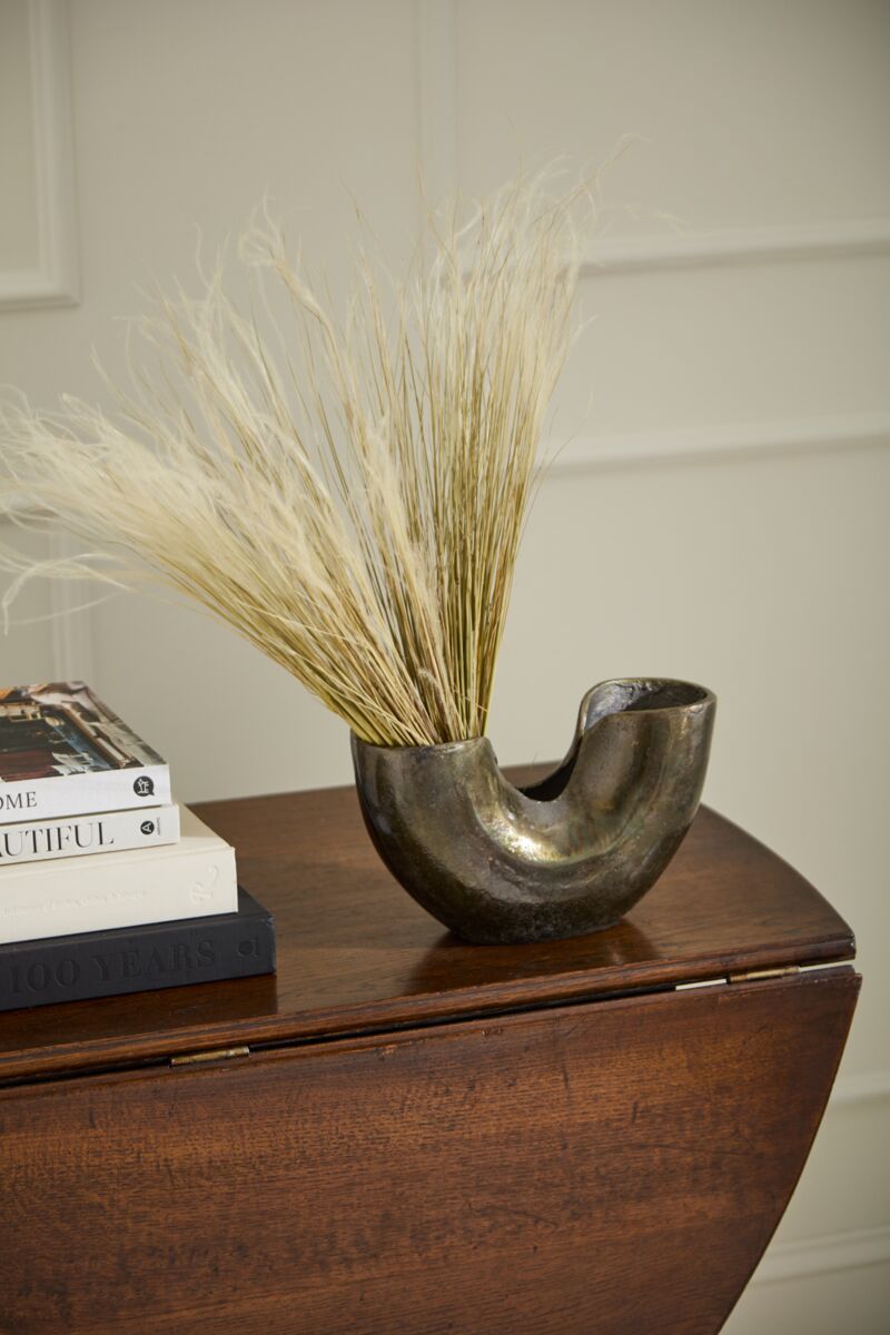 The Franz Vase by Accent Decor, crafted from recycled cast aluminum with a dark oxidized finish and filled with dried pampas grass, sits on a dark wooden table beside a stack of books against a light-colored wall.