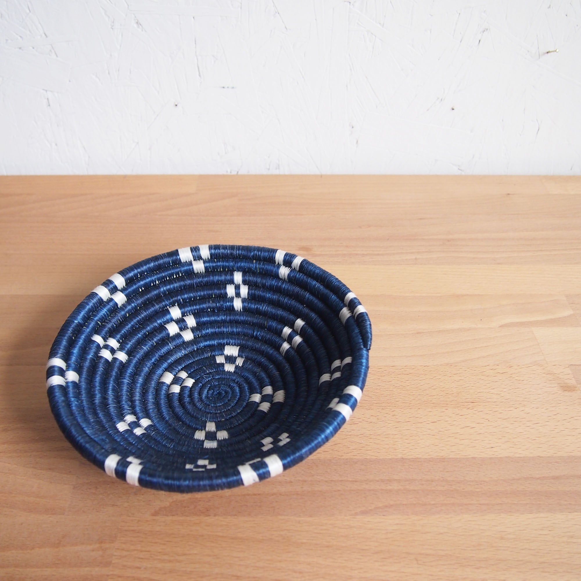 The Kabaya Small Bowl by Amsha - Faire, a round, shallow Rwanda basket with a blue and white geometric pattern, rests on a light wooden surface against a white textured wall.