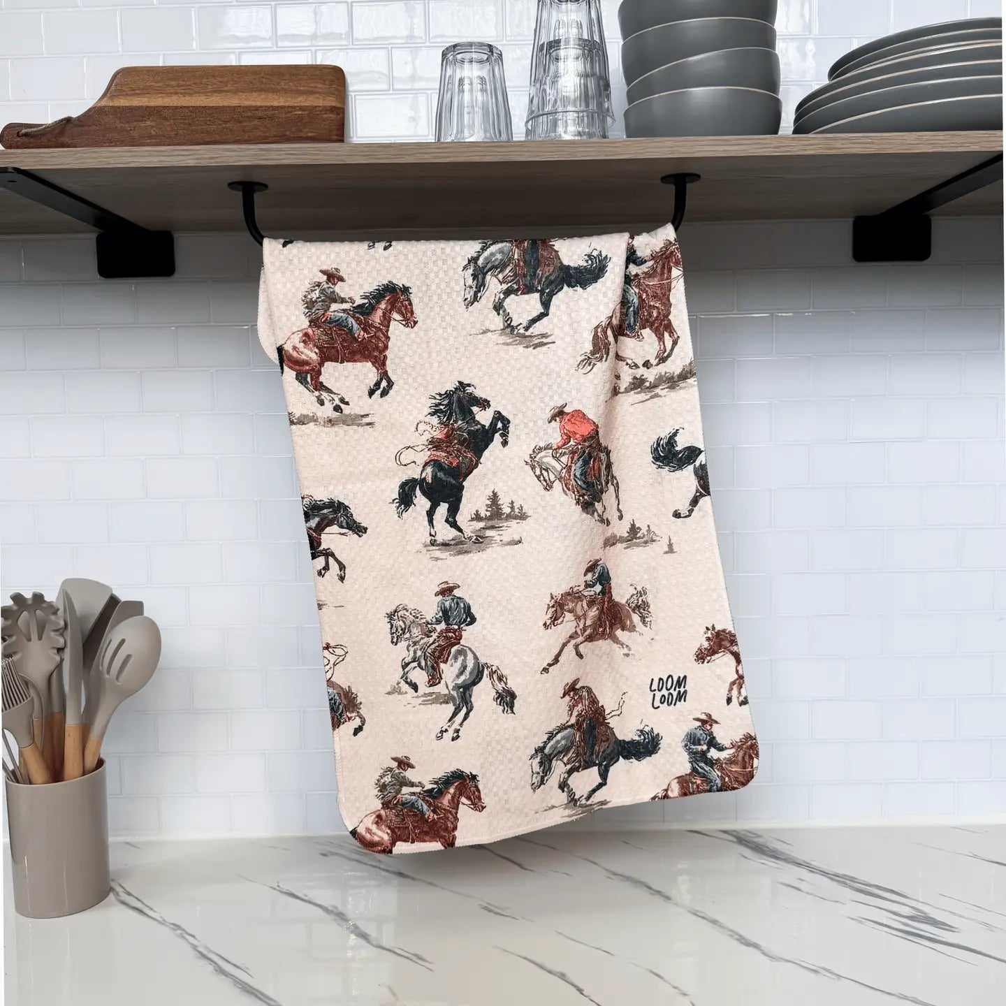 The Faire Rodeo Towel, featuring a cowboy-on-horseback pattern, hangs from a shelf below stacked dishes and glasses, above a countertop with a container of assorted kitchen utensils.
