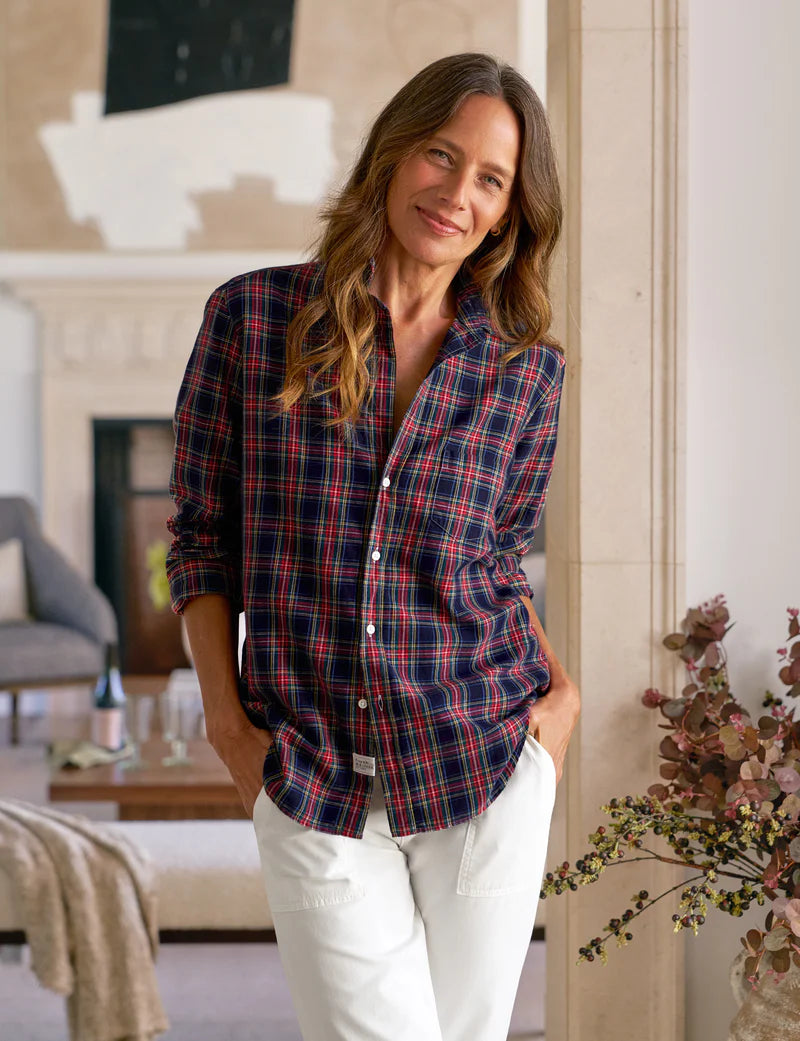 A woman with long brown hair stands indoors, smiling softly in the Frank & Eileen Eileen Button Up Shirt F25— a relaxed-fit red and navy plaid shirt with flattering button placement—paired with white pants, leaning against a beige wall.