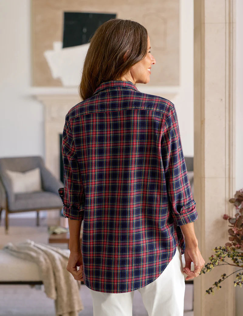 A woman with long brown hair stands indoors, facing away, wearing the Frank & Eileen Eileen Button Up Shirt F25 in red, blue, and white plaid with rolled-up sleeves and white pants. The room has neutral decor and artwork in the background.