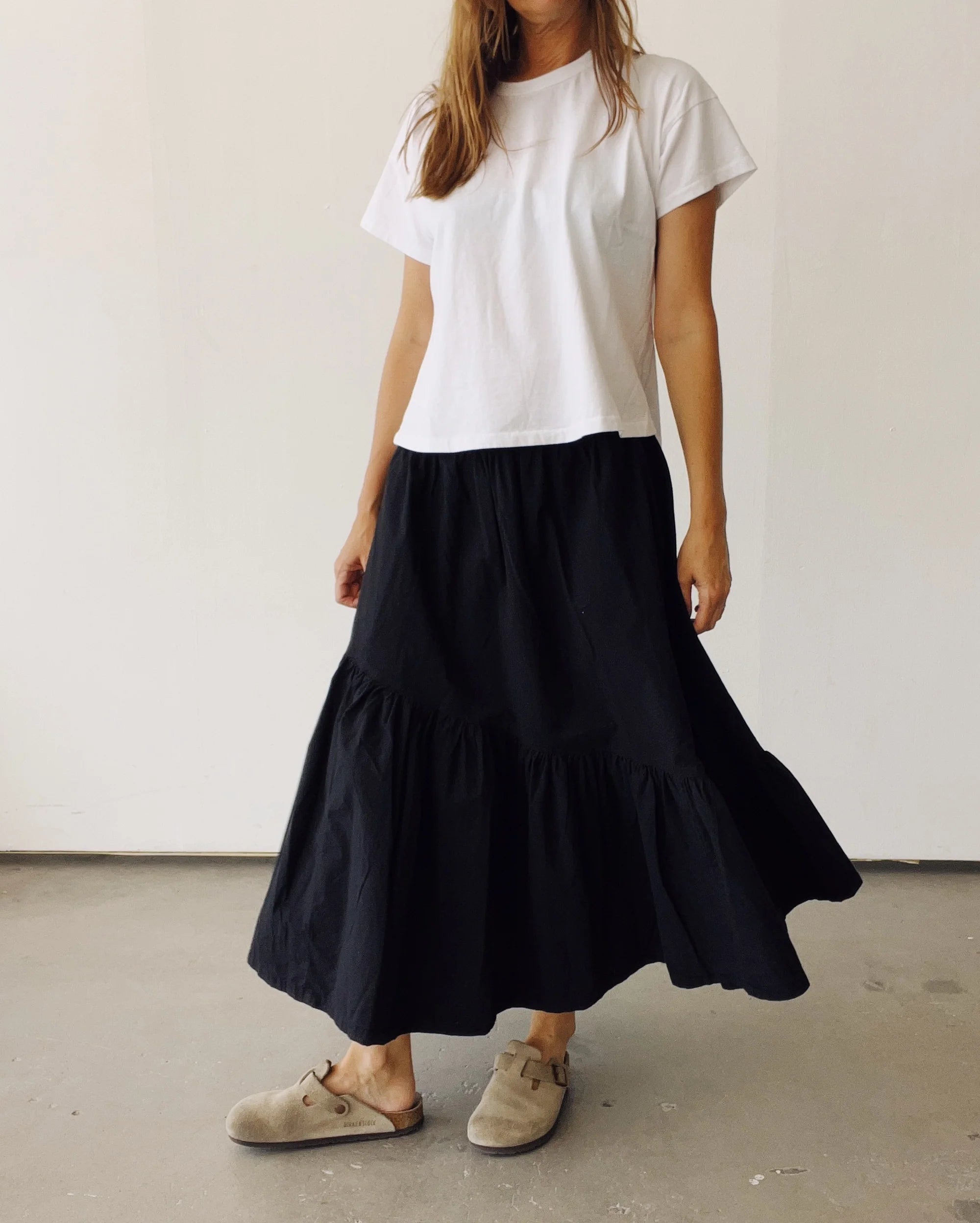 A woman stands indoors against a light wall, wearing Hey Gang’s The Ranch Skirt—a long black cotton skirt with an elastic waist—paired with a white short-sleeve t-shirt and beige clogs. Her face is not visible.