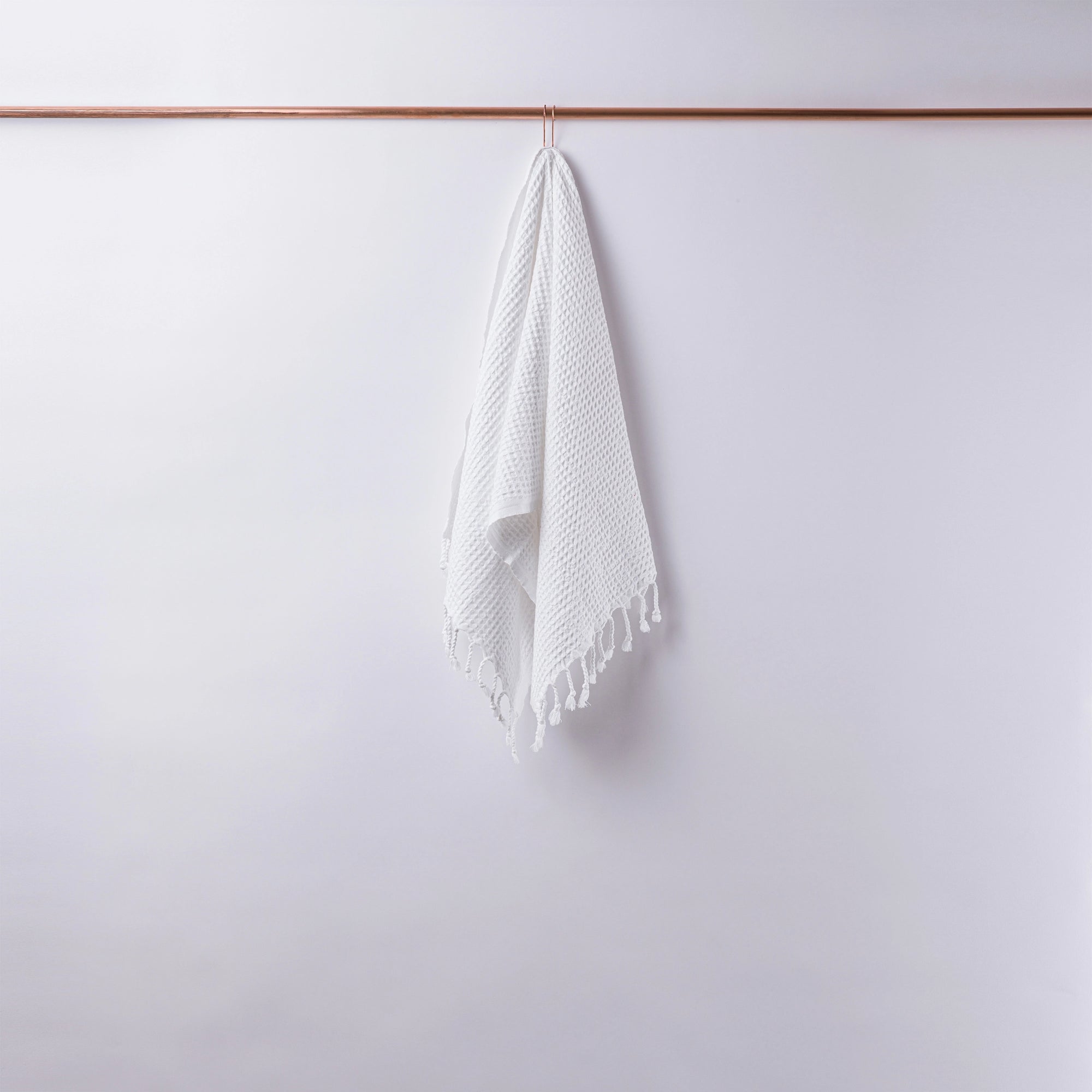 The Faire Waffle Hand Towel, featuring a white waffle weave and tassels, hangs neatly from a slim wooden rod against a light gray wall.