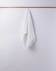 The Faire Waffle Hand Towel, featuring a white waffle weave and tassels, hangs neatly from a slim wooden rod against a light gray wall.