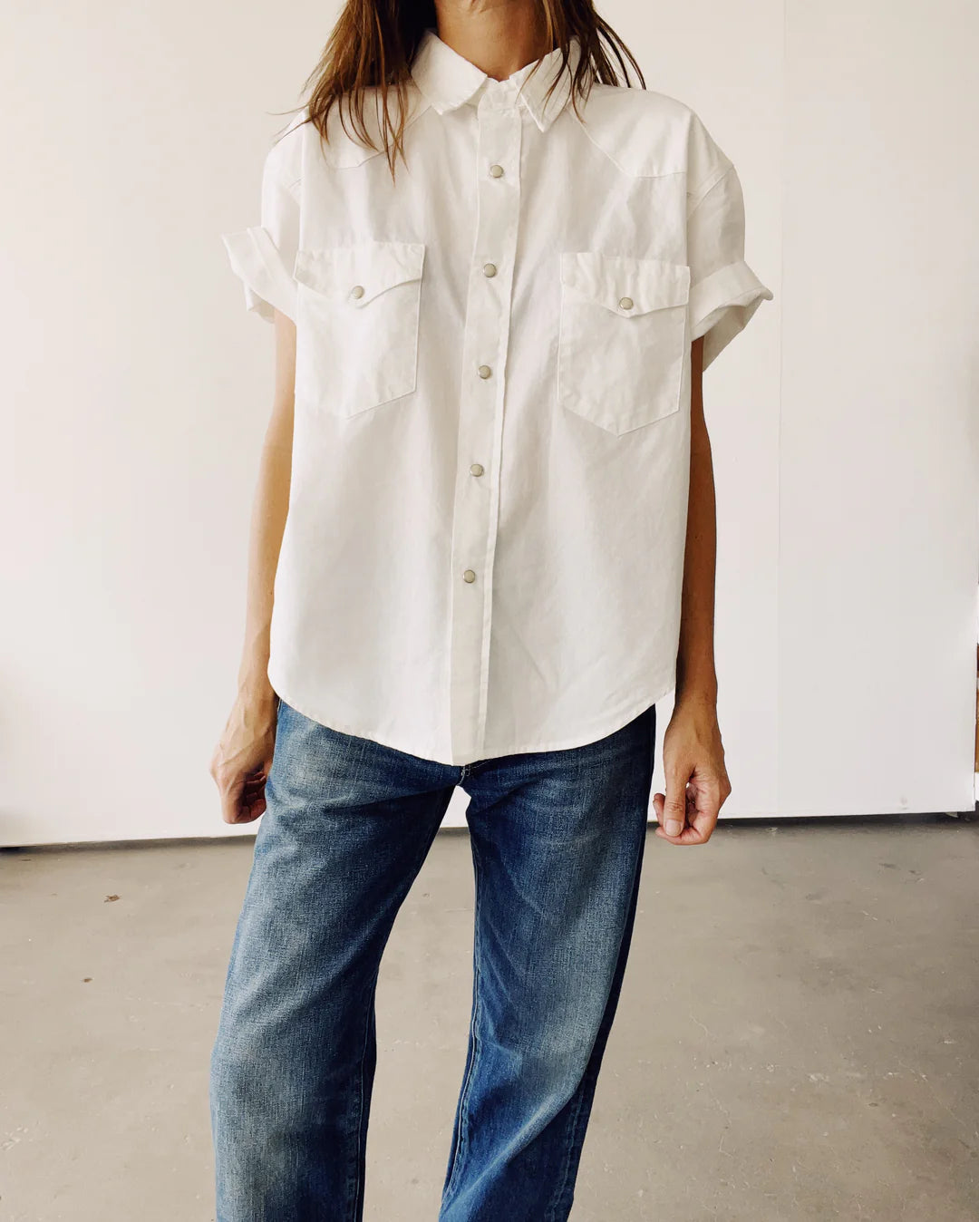 Someone is indoors wearing The Western Shirt, an oversized short-sleeve button-up with two chest pockets from Hey Gang, paired with blue jeans. The background maintains a minimal and neutral aesthetic.