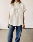 Someone is indoors wearing The Western Shirt, an oversized short-sleeve button-up with two chest pockets from Hey Gang, paired with blue jeans. The background maintains a minimal and neutral aesthetic.