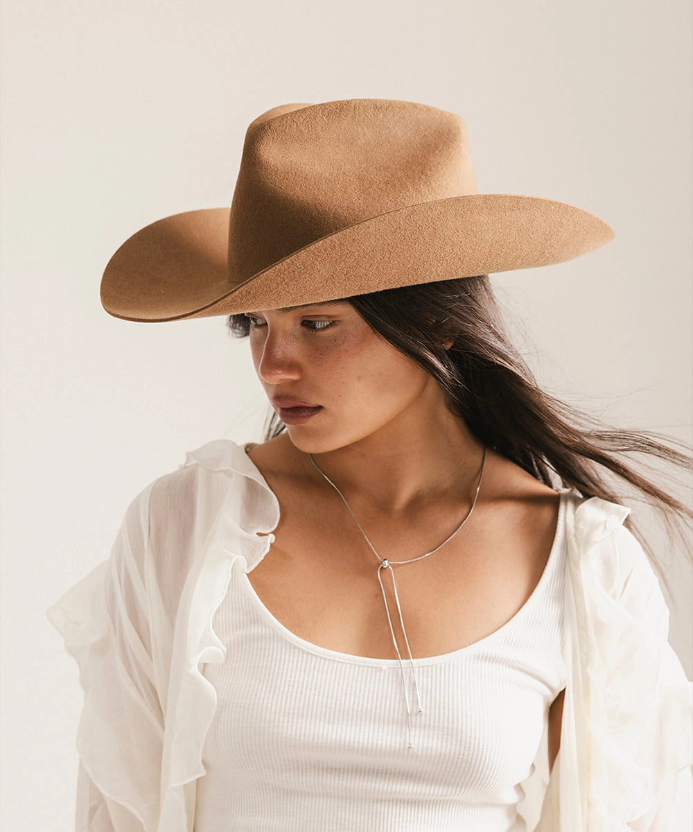 A woman wearing the Faire Charlie Western Hat and a white tank top with a sheer ruffled shirt looks downward, her long hair over one shoulder and a minimalist silver necklace completing her look.
