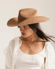 A woman wearing the Faire Charlie Western Hat and a white tank top with a sheer ruffled shirt looks downward, her long hair over one shoulder and a minimalist silver necklace completing her look.