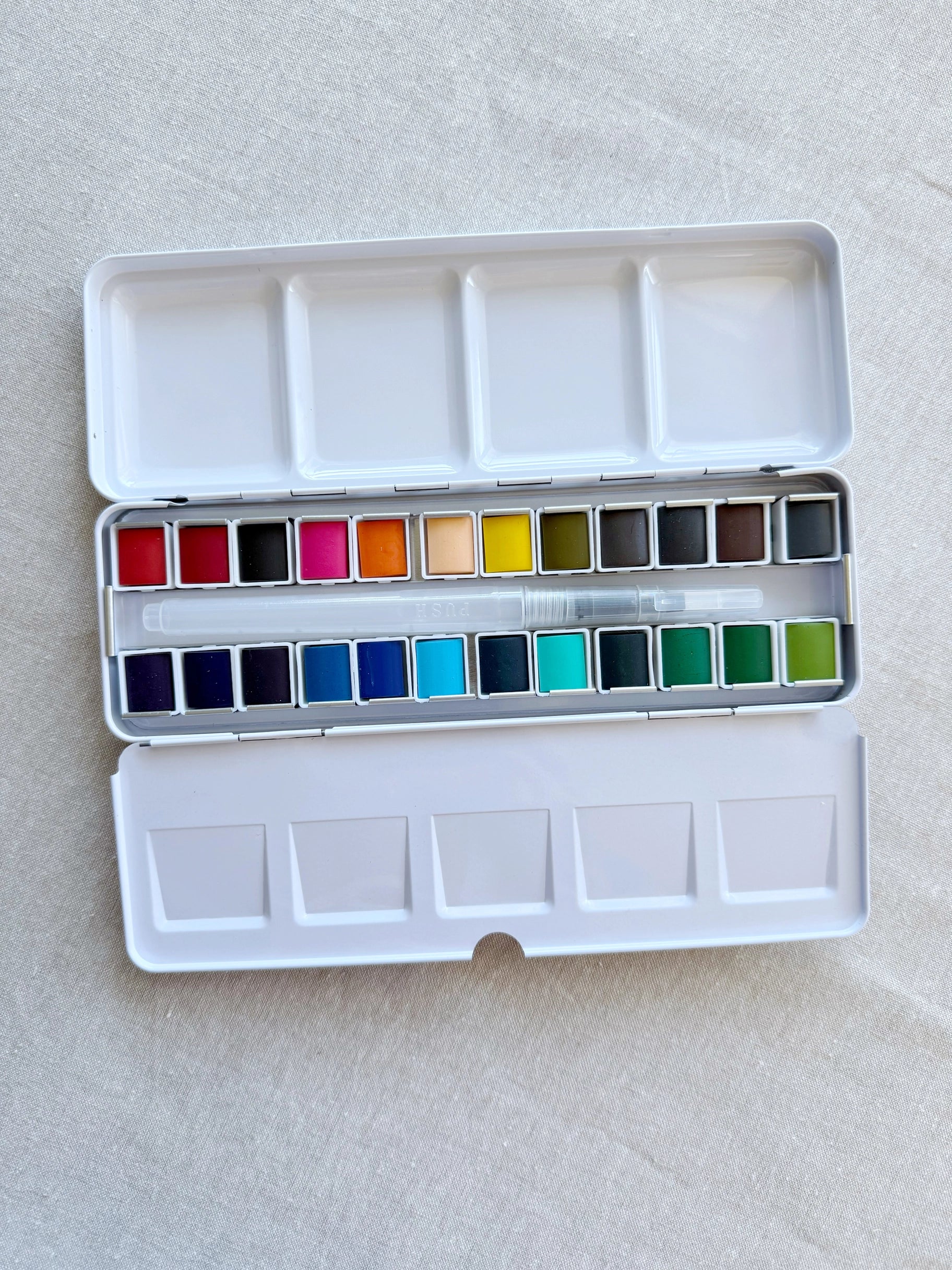 The Faire Watercolor Paint Set w/ Brush features 24 assorted color pans in a white plastic tin, with empty mixing wells on the lid and base, shown on light fabric.